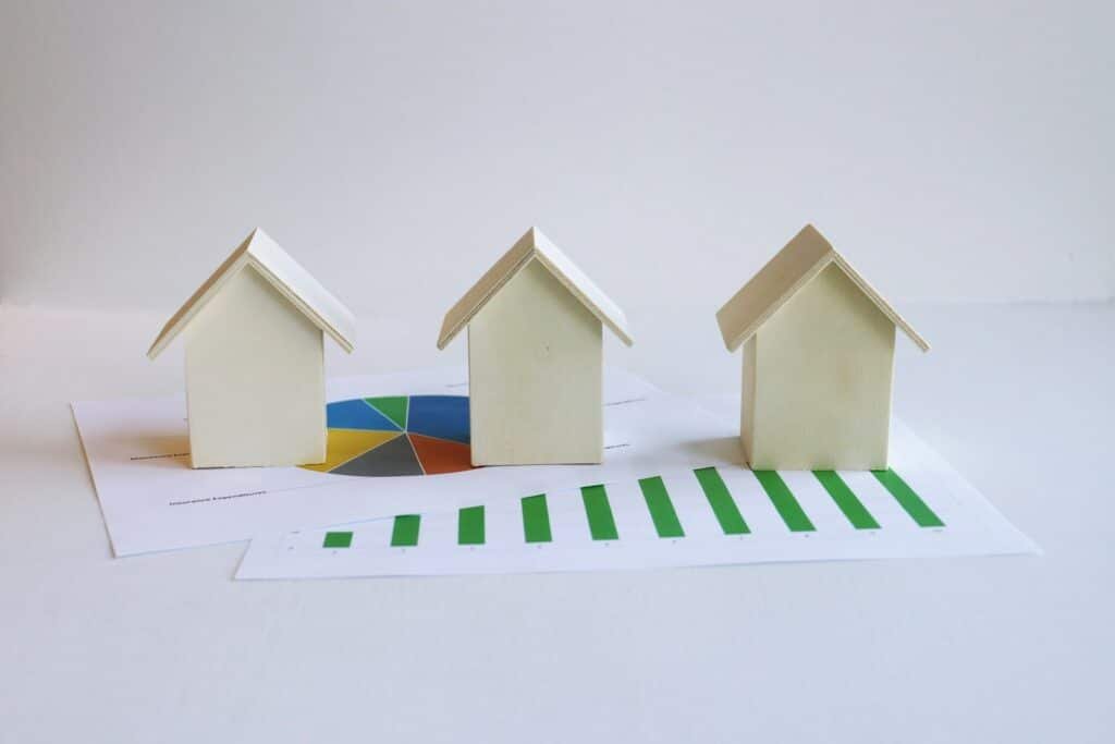 Three small wooden house models placed on top of printed property data charts, including a pie chart and a bar graph, against a white background