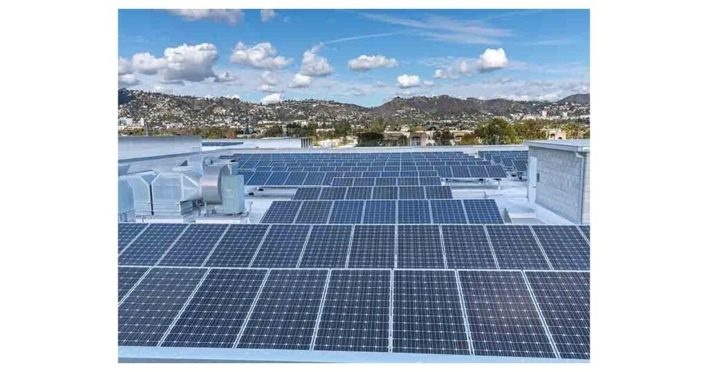 Solar panels installed on a flat commercial building rooftop, with ventilation equipment visible, hills and residential buildings in the background under a partly cloudy sky