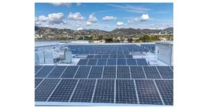Solar panels installed on a flat commercial building rooftop, with ventilation equipment visible, hills and residential buildings in the background under a partly cloudy sky